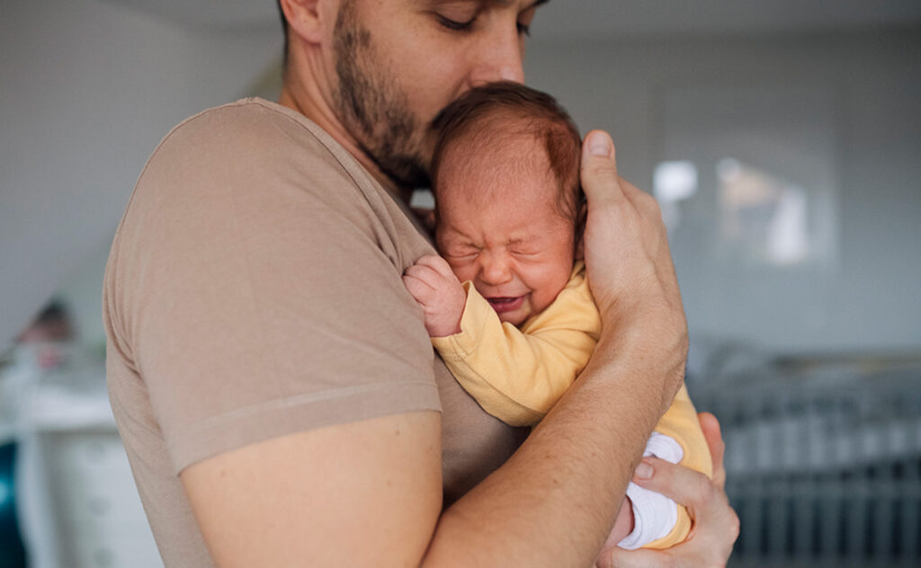 Unrecognizable father holding his crying baby girl in hands and kissing her
