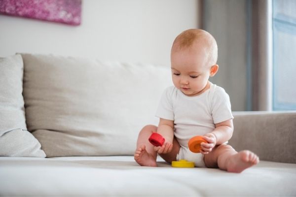 Baby with blond hair sitting up on bed playing with coloured blocks