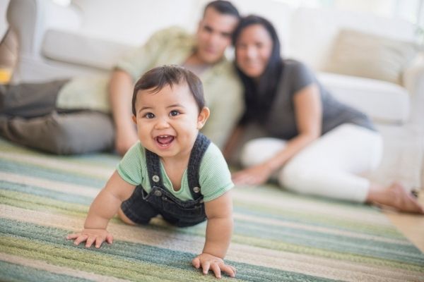 Smiling dark haired toddler boy crawls away from his parents in a lounge room