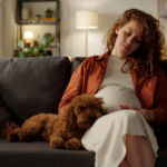 Woman with long red hair, 19 weeks pregnant, sitting on sofa with dog cuddled up