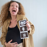 Smiling pregnant woman with curly hair clutching her baby bump while holding up a sheet of ultrasound images