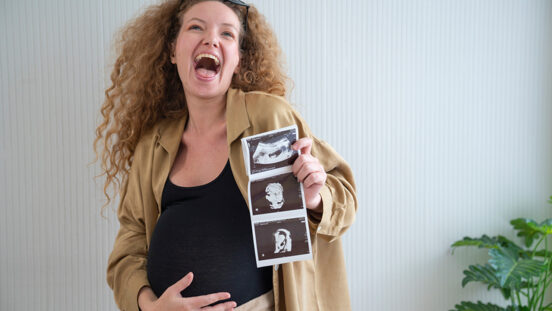 Smiling pregnant woman with curly hair clutching her baby bump while holding up a sheet of ultrasound images