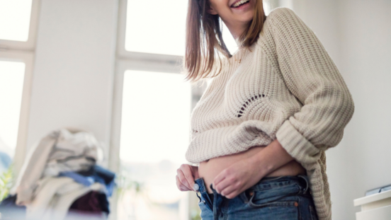 Pregnant woman smiling as she tries to do up jeans