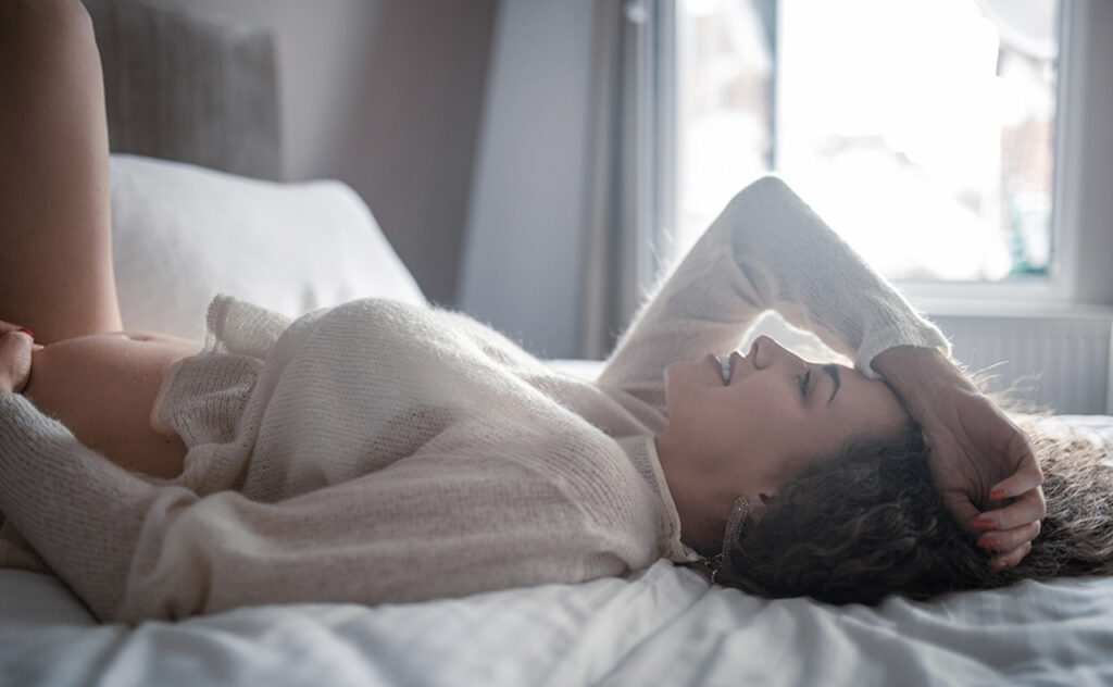 Beautiful young pregnant woman is touching pregnant belly while lying on the bed in bedroom and enjoying morning lights