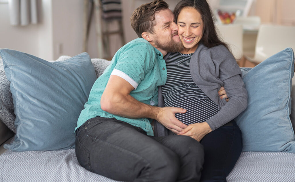 A mixed race couple is snuggling on their couch at home. The woman of Asian and Pacific Islander descent is pregnant with their first child. She is in her second trimester. The woman's husband is kissing her cheek and holding her belly. The happy soon to be parents are excited for the many adventures parenthood will bring.