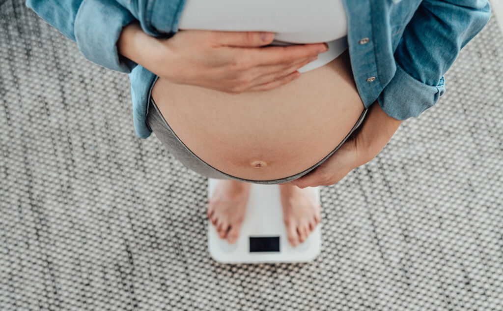 High angle view of pregnant woman touching her baby bump while standing on weight scale. Healthy pregnancy.
