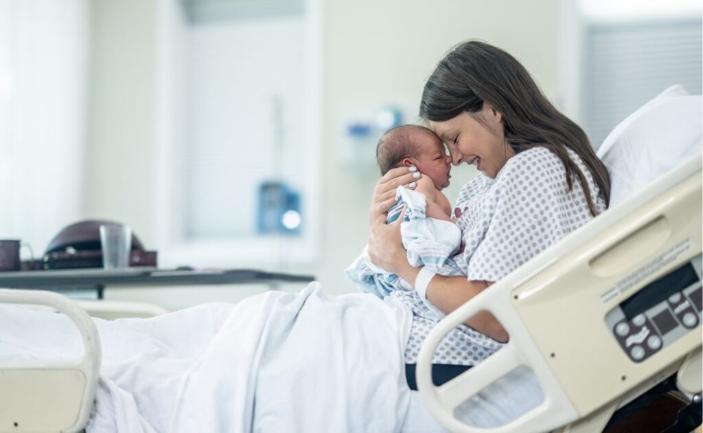 A new Mother sits up in her hospital bed shortly after delivery as she holds her newborn out in front of her and studies his features. She is wearing a hospital gown and is laying in her hospital bed with the inafnt.
