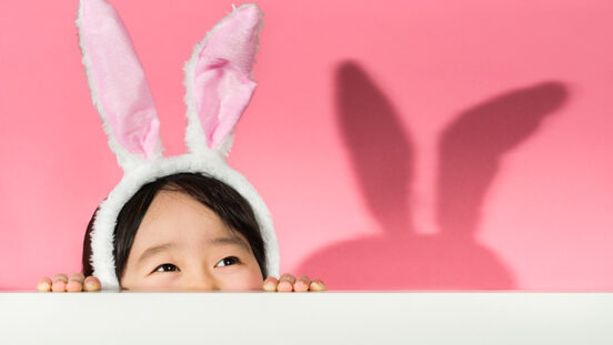 Young dark haired girl wearing bunny ears peering over wall with a pink background.