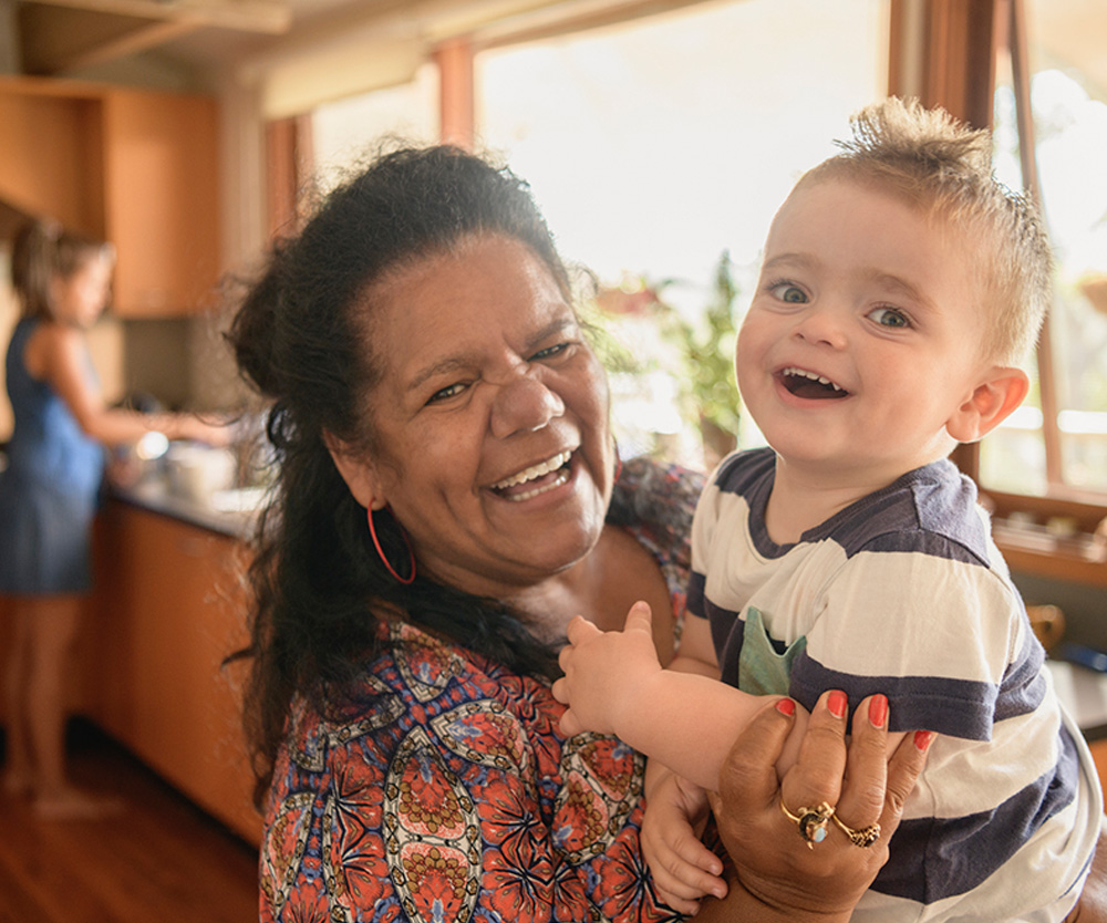 Portrait of Aboriginal grandmother holding baby grandson. Young boy and mature woman smiling towards camera, mother in background doing dishes