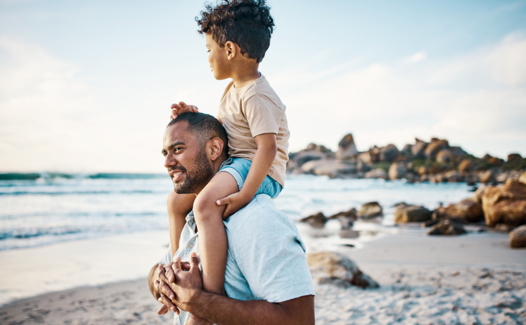 Dark haired dad on the beach with his dark hair young son on shoulders