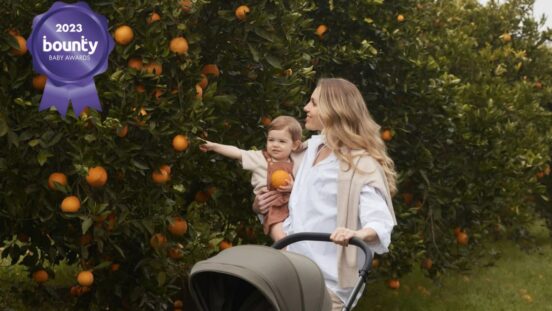 Woman with long blond hair pushing a stroller and carrying a toddler on her hip as they walk by an orange bush