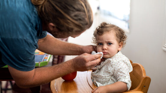 Dad wipes his toddler's face while she sits in her high chair