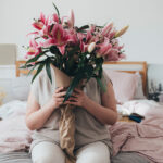 An overweight woman sitting on her bed and holding a bouquet of pink lillies