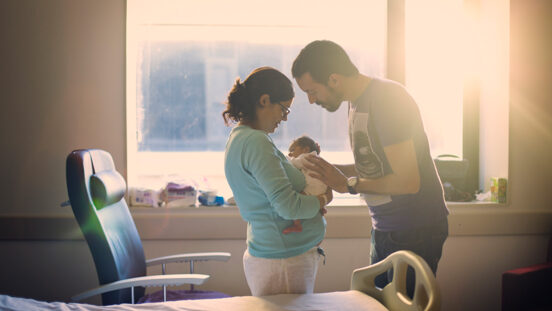 New parents stand in front of sun streamed hospital room window while staring at their newborn.