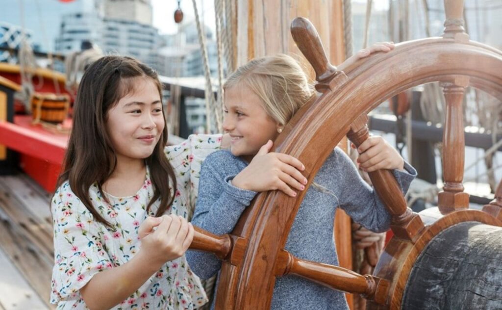 Two tween girls holding on to a ship's steering wheel at the Australian National Maritime Museum 

