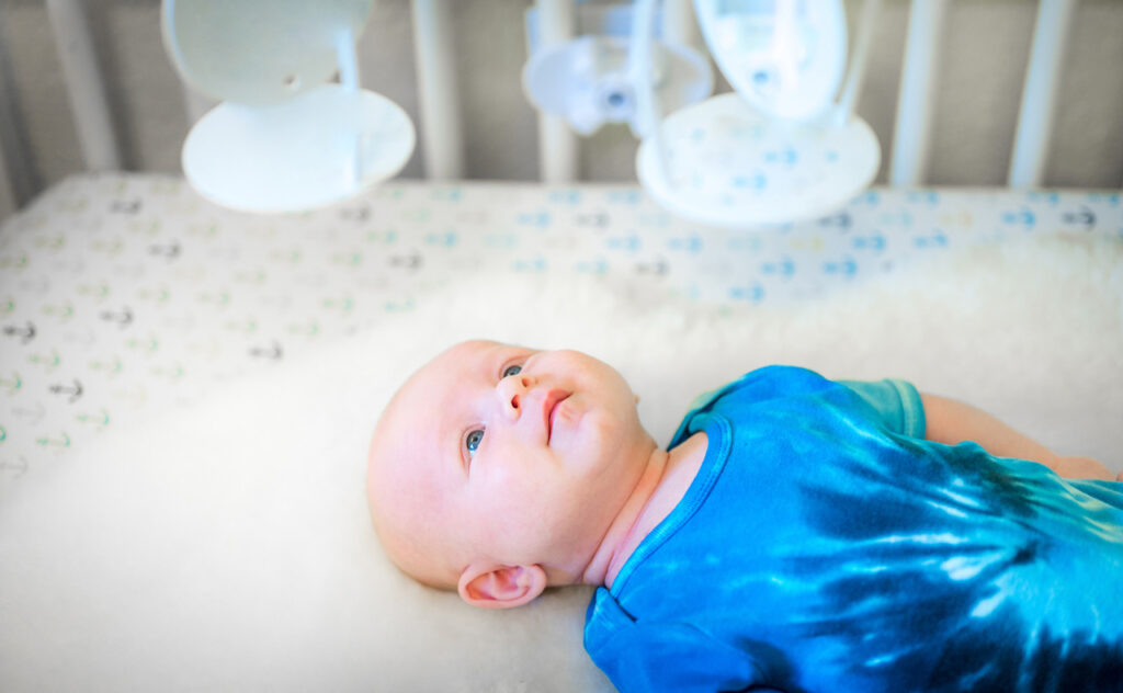 newborn infant in tided onesie laying on crib staring up at mobile.