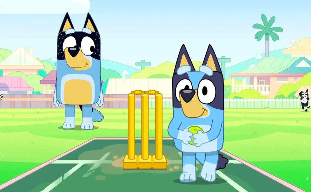 A still image of an ABC TV Bluey episode showing Bluey and Bandit on a cricket pitch, Bandit standing behind the wickets while Bluey prepares to bowl.