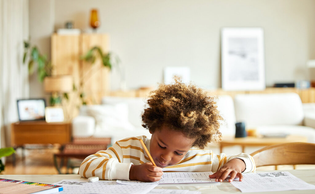 African American boy with pencil writing on paper at table. Male is doing homework. He is wearing casuals at home.