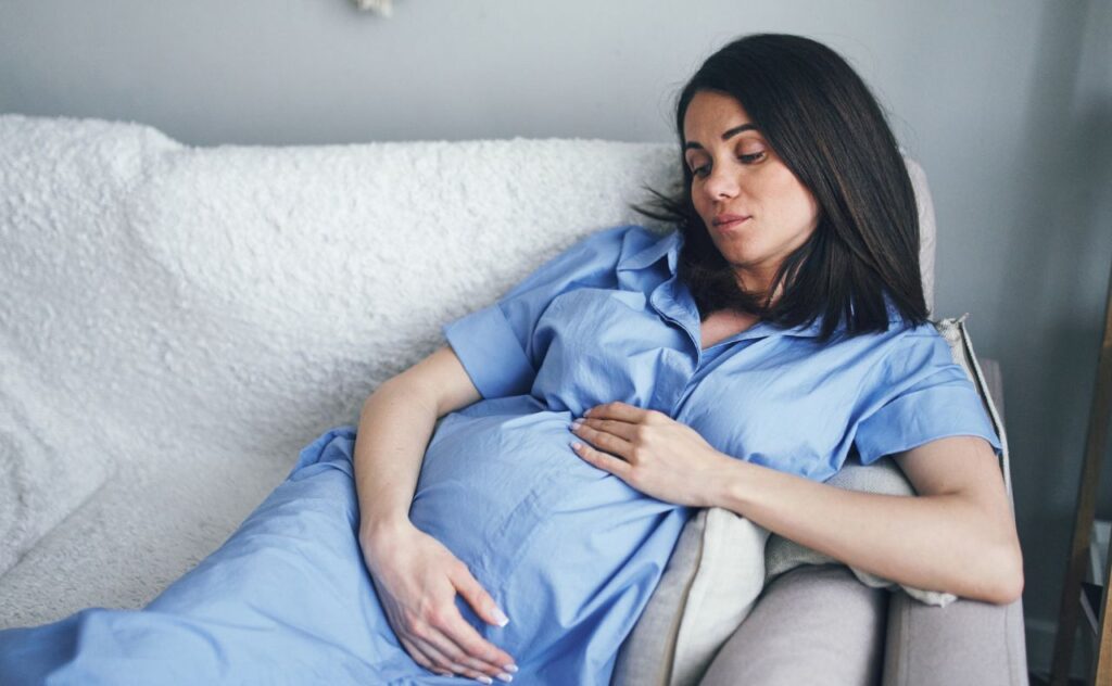 Women with shoulder length black hair wearing a long pale blue shirt dress and cradling her baby bump