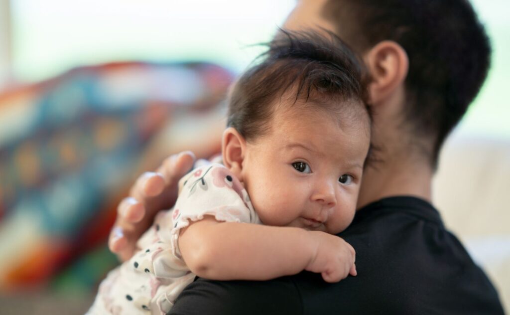 Close up of a baby with dark hair looking at the camera over her dad's shoulder