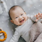 Smiling newborn looking and reaching up to camera while laying on grey blanket.