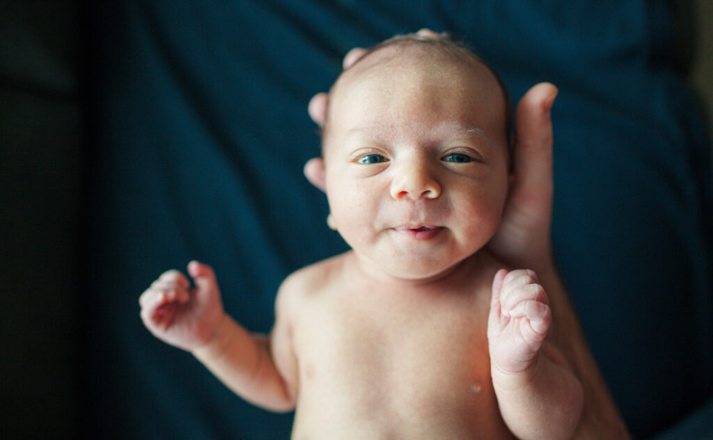 Newborn baby, head resting in adults hand staring up at camera.