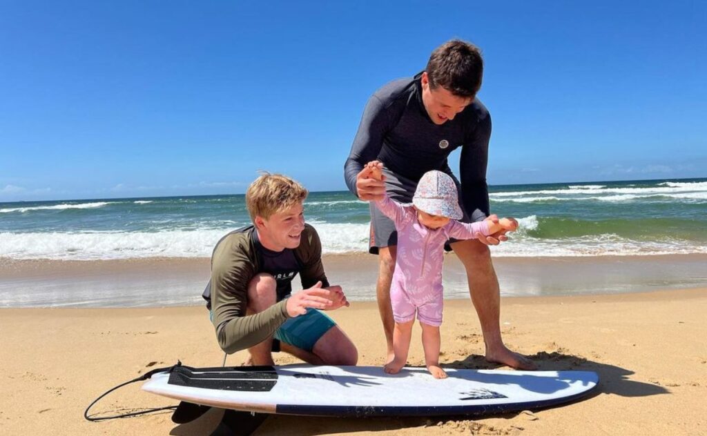 Baby Grace being held standing up on a surfboard while uncle Robert smiles