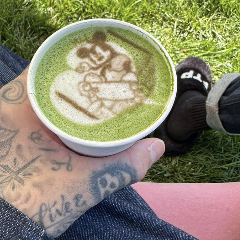 Travis barker's tattooed hand holding a cup of green matcha drink with a Mickey Mouse art scene in the froth.