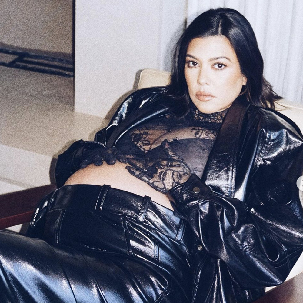 Pregnant Kourtney Kardashian Barker reclining, holding her stomach while wearing black leather pants, black leather jacket and black lace crop top.