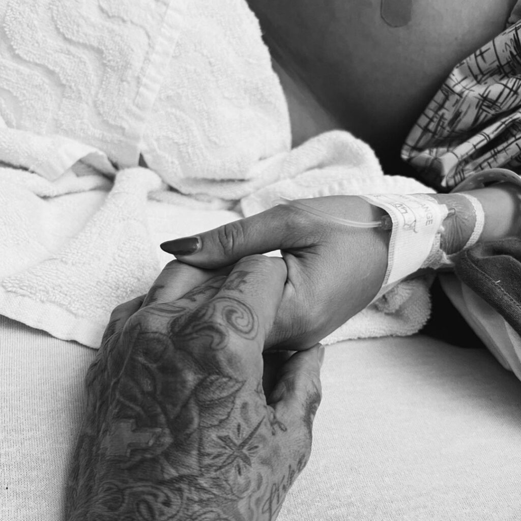 Black and white image of Travis Barker's tattooed hand holding wife, Kourtney Kardashian Barker's hand on hospital bed.