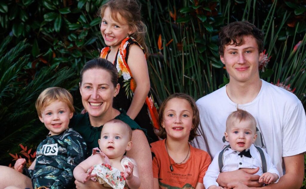 Jana and all five kids sitting together, smilng at camera in an outdoor setting