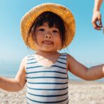 LIttle girl with dark hair and a straw hat, wearing blue and white striped swimmers at the beach