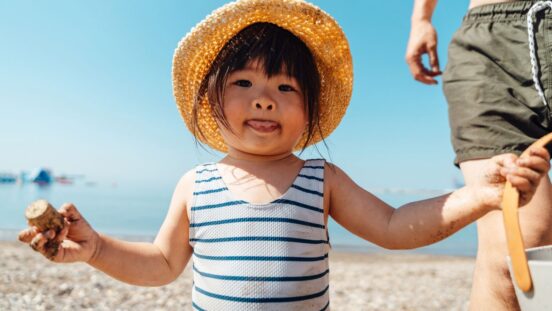 LIttle girl with dark hair and a straw hat, wearing blue and white striped swimmers at the beach