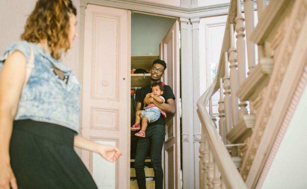 Fair-haired woman with pale blue top and dark skirt looking back at dark-haired man standing in a doorway holding a toddler girl