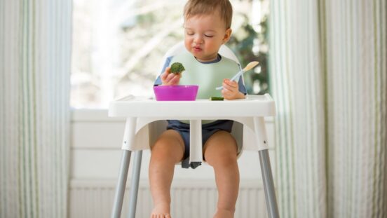 Toddler boy sits in highchair in front of a window looking at broccoli