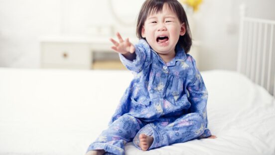 Dark haired toddler in blue pyjamas sitting on a bed, crying and reaching out with one hand