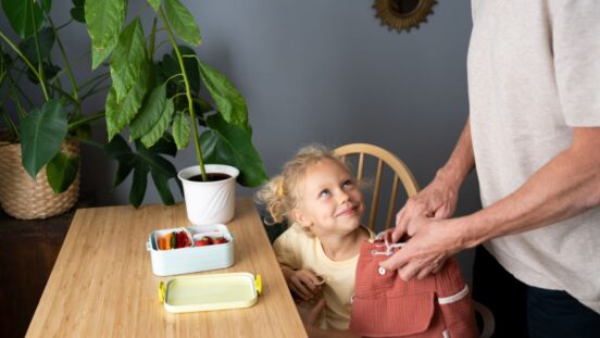 Little girl with blond ponytail sitting a table with a vegetarian meal on it looking up at a parent packing their bag