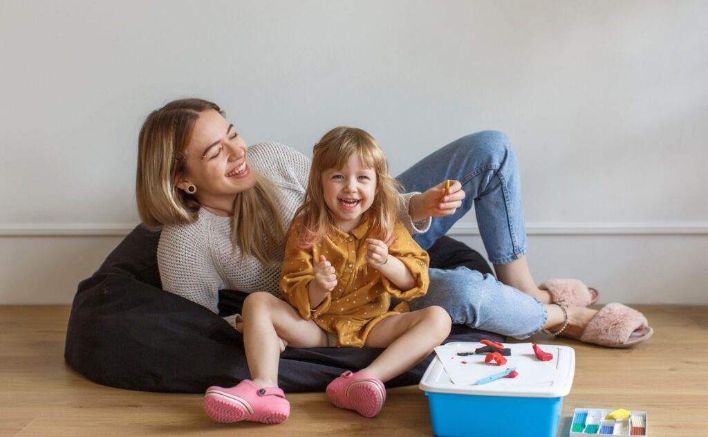 Laughging young woman with long blonde hair sits on a black beanbag on the floor with a laughing toddler girl looking at the camera