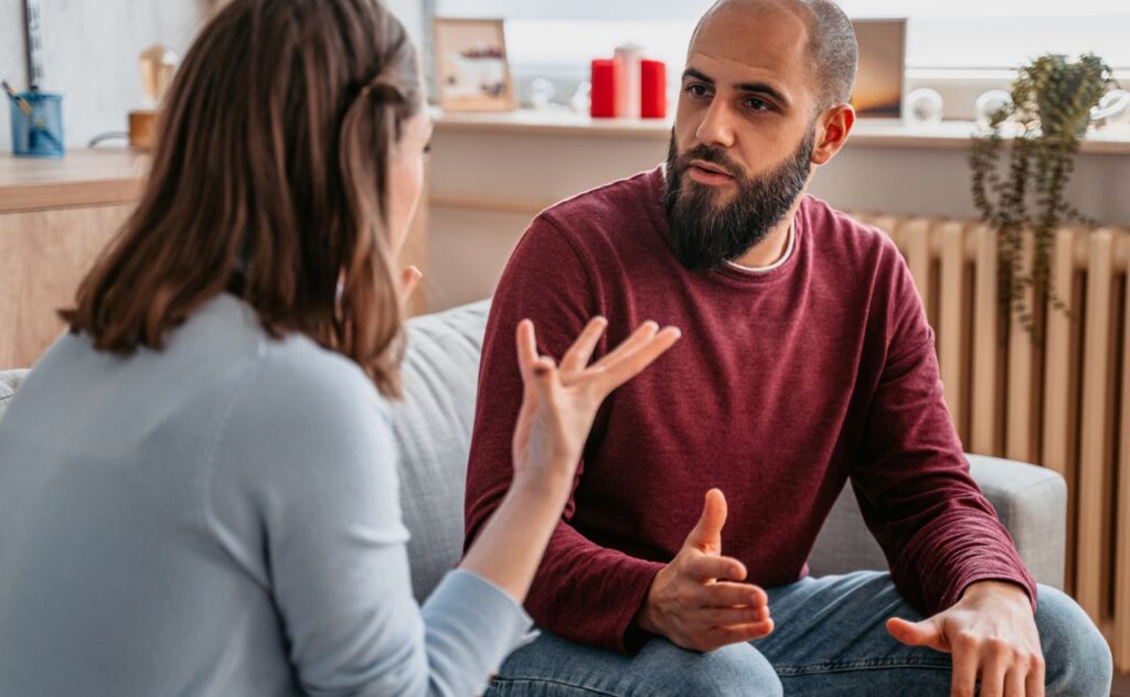 Man with dark beard and receeding hairline sits on a sofa seeming to argue with a woman with dark shoulder-length hair holds her hands up