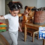 Toddler girl with dark hair in bunches walks through a play room holding an adult's hand