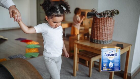 Toddler girl with dark hair in bunches walks through a play room holding an adult's hand