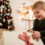 Caucasian toddler boy sitting near Christmas tree, and unwrapping his Christmas present