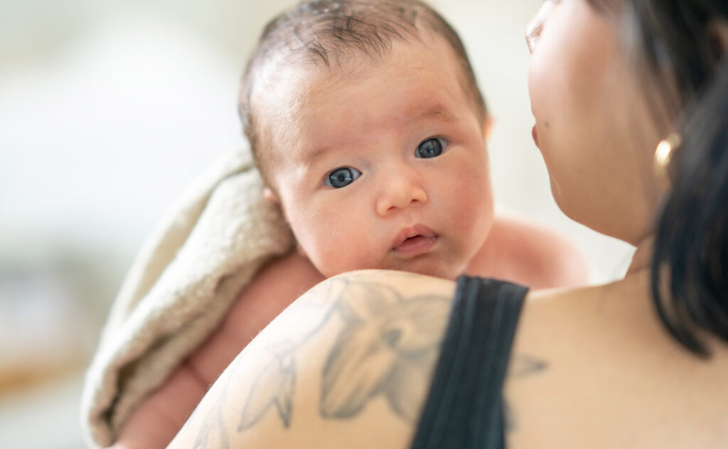 A young mother carries her baby on her shoulder as she walks around the nursery and burps her child after feeding him. The baby is skin to skin with his mother and lightly wrapped in a blanket over it.