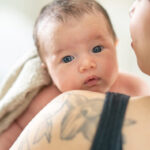 A new mother holds her infant up over her shoulder as she walks around the nursery and burps her child after feeding. The baby is skin-to-skin with her mother and lighting wrapped wth a blanket overtop.