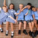 Five school friends in school uniform sitting on brick wall pulling faces and smiling towards camera