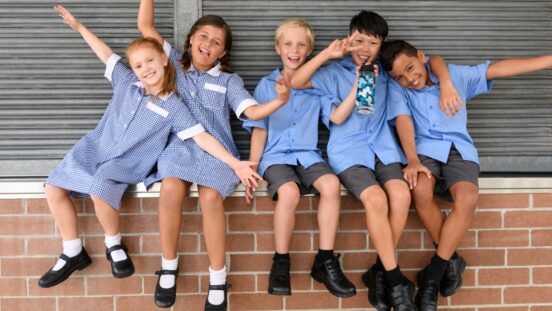 Five school friends in school uniform sitting on brick wall pulling faces and smiling towards camera