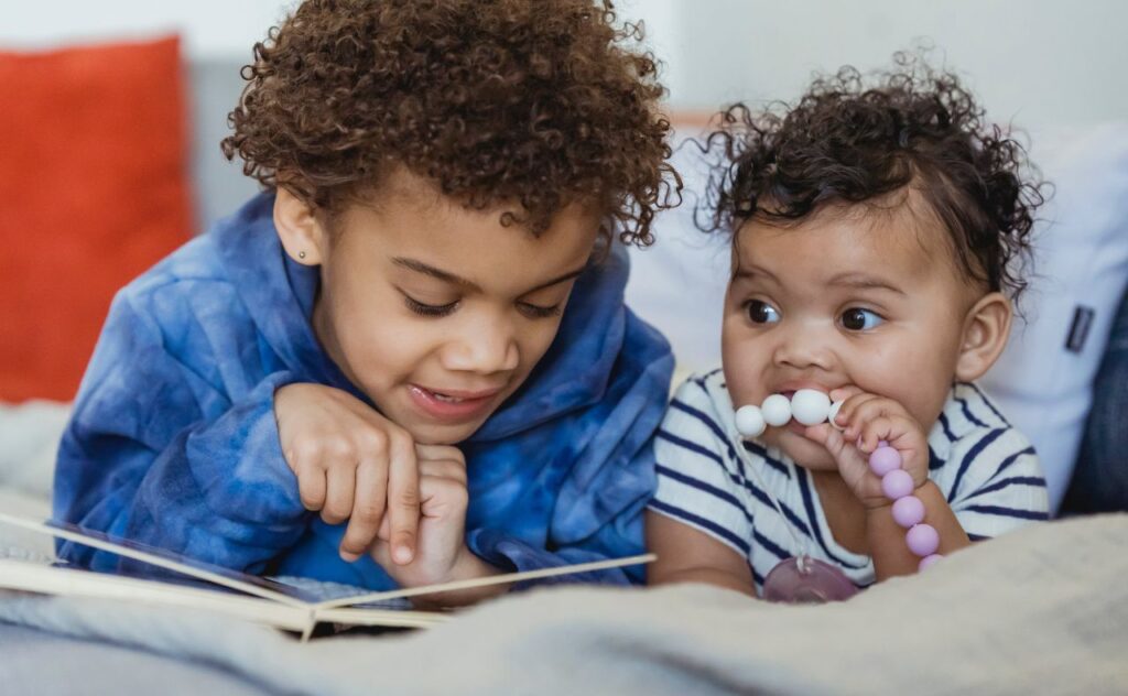 Dark hair preschooler lying on tummy and pointing at book as they read out loud to their little sibling