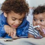 Dark hair preschooler lying on tummy and pointing at book as they read out loud to their little sibling