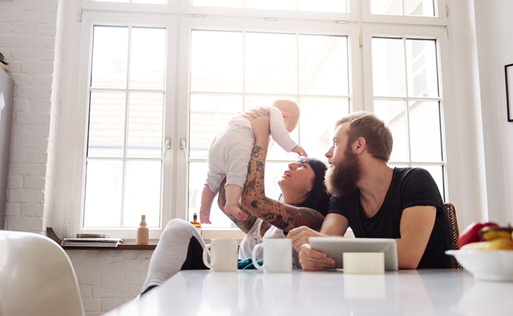 Young tattoed mother and father with newborn baby sitting in their kitchen and having fun together