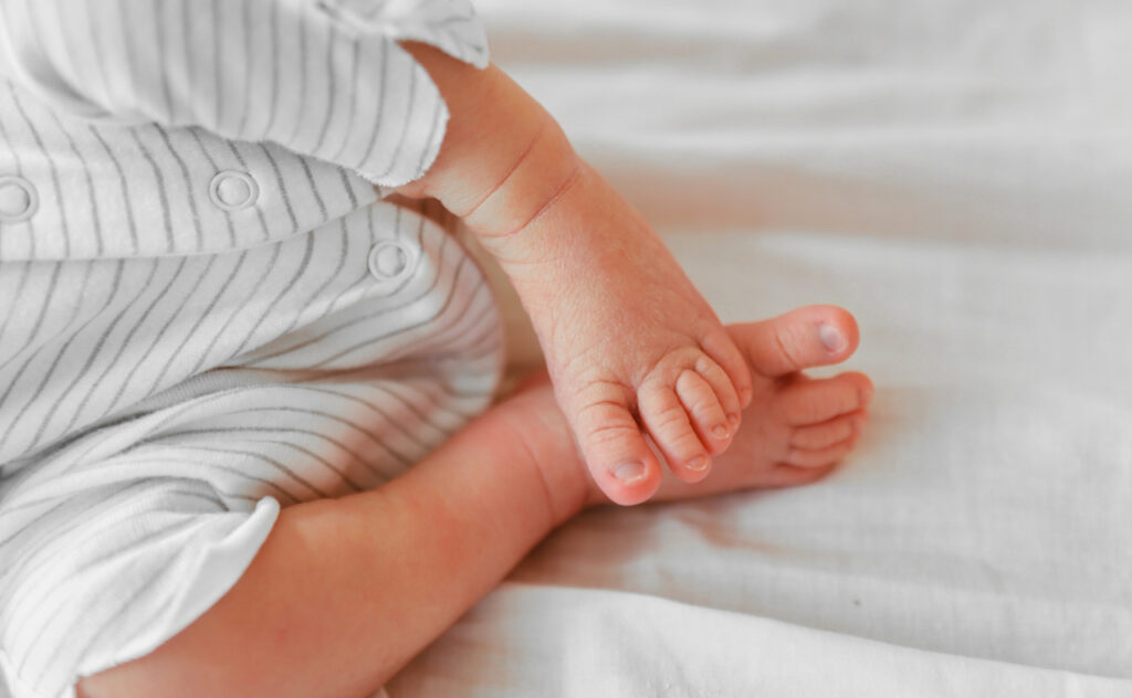 Cute sleeping newborn baby feet on white blanket. Baby in a white gender neutral romper on the bed. Little toes, heels.